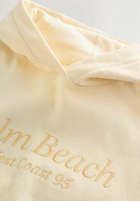 Gaiši dzeltena kapučjakas ar izšūtu uzrakstu "Palm Beach West Coast 95" uz krūtīm, izšūtā dzijas krāsā.