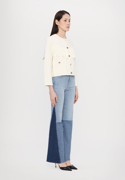 Weißer, fransiger Button-Up-Jacke kombiniert mit hellblauen, ausgestellt geschnittenen Jeans, die dunkelblaue Besatz-Akzente aufweisen, und spitzen schwarzen Absätzen.