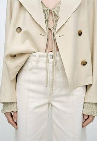 Beige double-breasted jacket over a textured olive top, paired with high-waisted white denim pants featuring visible stitching and a button.