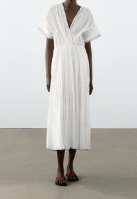 Personne à la peau foncée portant une robe midi blanche plissée à manches courtes avec des bordures noires et des sandales rouges, debout sur un sol gris.