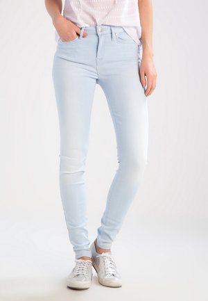 Femme portant un jean skinny bleu clair et des baskets blanches, debout avec une main dans la poche sur un fond uni.