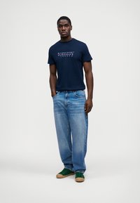 Ναυτικό μπλε βαμβακερό t-shirt με λογότυπο "tommy jeans" σε κόκκινο και λευκό χρώμα; ανοιχτό μπλε τζιν παντελόνι; πράσινα αθλητικά παπούτσια με καφέ σόλες.