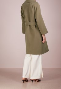 Manteau en mélange de laine vert olive avec des manches larges, taille ceinturée et fente au dos. Porté par-dessus un pantalon large blanc et des talons noirs.