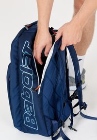 Marineblauwe rugzak met gequilt ontwerp, grote witte rits en opvallend 'Babolat' logo in lichtblauw. Verstelbare schouderbanden en oranje hardware accenten.