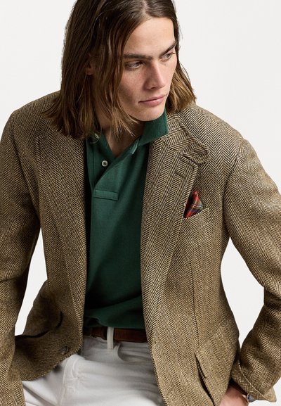 Sacou maro cu model herringbone, cu umeri, purtat peste o cămașă polo verde. Buzunarul are un batistă cu un model în carouri roșii și albastre. Pantaloni albi.