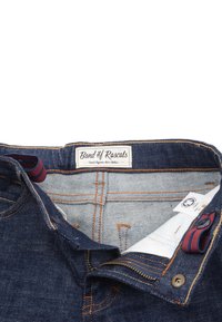 Lasten farkut tummansinisellä pesulla, kontrastivärisellä oranssilla tikkaus ja punaruudullinen sisävyö. Tagi lukee &quot;Raitapojat.&quot;