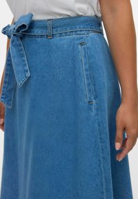 Gonna di jeans azzurro chiaro con cintura a fiocco, tasche laterali, forma a trapezio, dettagli di cucitura visibili e una texture liscia.