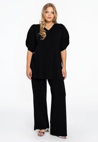 Schwarzes Oberteil mit Puffärmeln und V-Ausschnitt, kombiniert mit weiten schwarzen Hosen. Das Outfit zeichnet sich durch eine glatte Textur und ein lässiges Design aus.