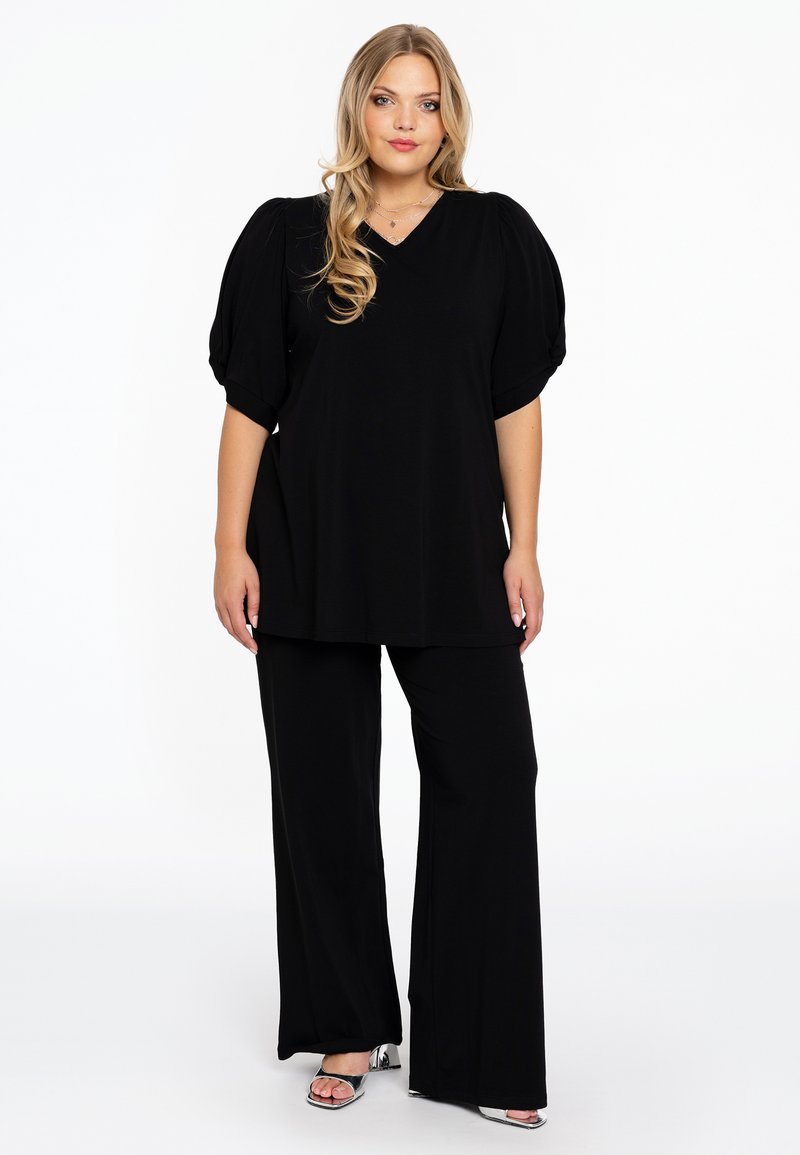 Schwarzes Oberteil mit Puffärmeln und V-Ausschnitt, kombiniert mit weiten schwarzen Hosen. Das Outfit zeichnet sich durch eine glatte Textur und ein lässiges Design aus.