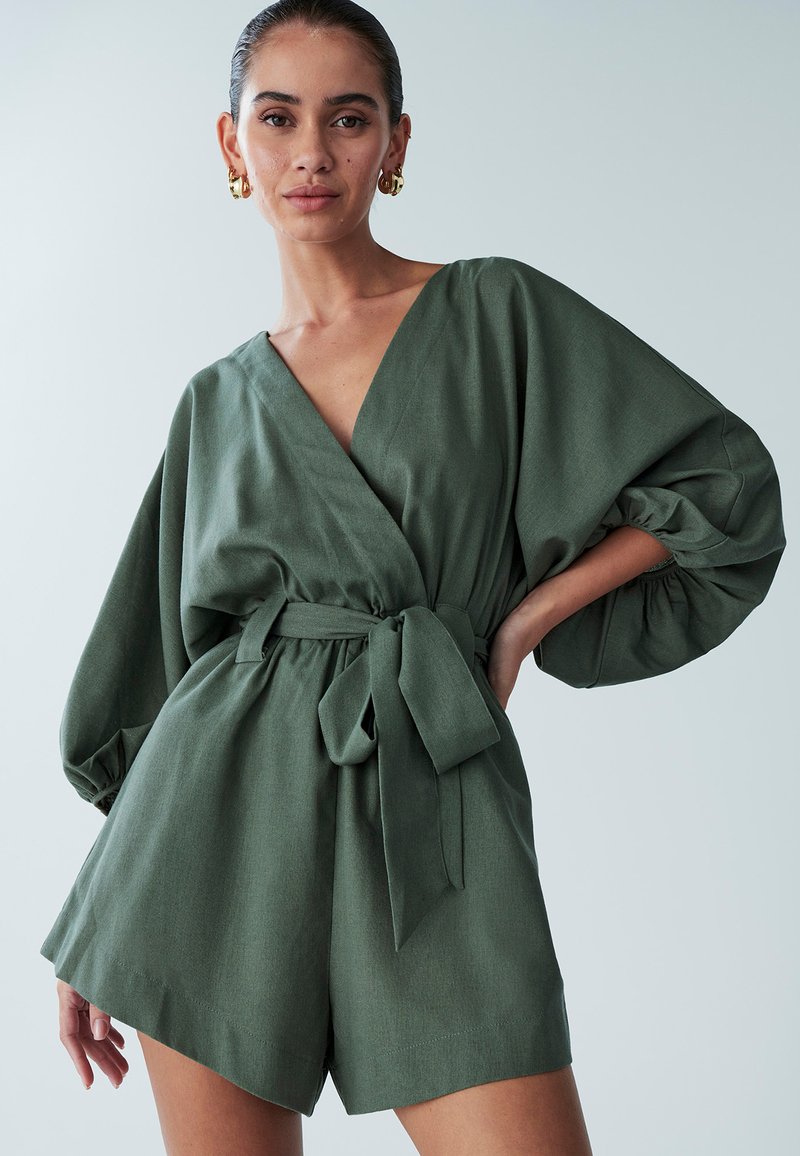 Olivovo zelený overal áčkového štýlu s širokými nadýchanými rukávmi a viazaním v páse. Vyrobený z textúrovanej látky, má šortky so strihom rovného strihu.