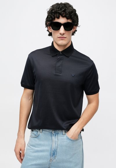 Melnas polo krekls ar apkakles kakla izgriezumu un īsām piedurknēm, ar nelielu logotipu un izgatavots no gludas auduma; kopā ar gaiši ziliem džinsiem.