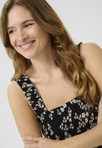 Robe noire à fleurs avec un corsage smocké et des bretelles larges, ornée de petites fleurs blanches et rouges. Cheveux coiffés en vagues lâches. Teint clair.