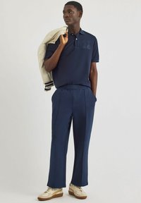 Marineblaues Poloshirt mit gesticktem Logo, kombiniert mit passenden Hose mit elastischem Bund und cremigen Sneakers, hält eine leichte Jacke über der Schulter.