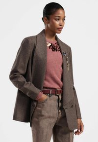 Blazer marrone a quadri houndstooth con rever strutturati, abbinato a un maglione rosa e jeans beige sbiaditi a vita alta, accentuato da una cintura marrone.