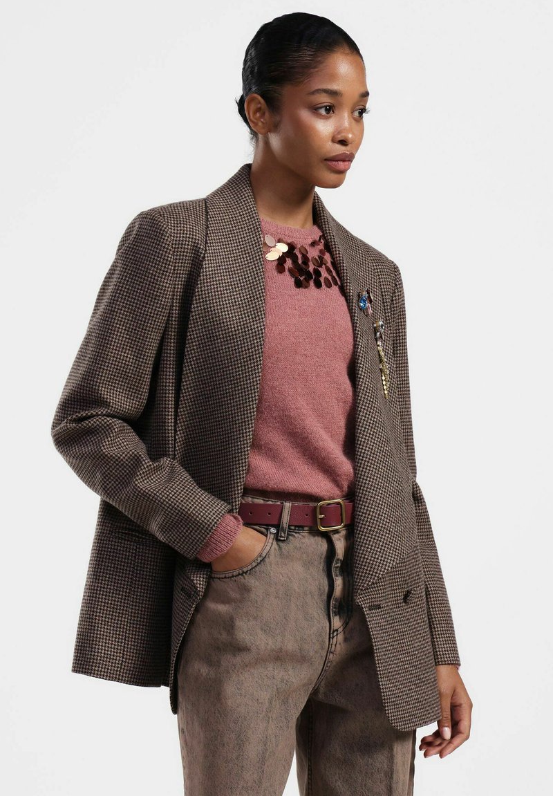 Blazer marrone a quadri houndstooth con rever strutturati, abbinato a un maglione rosa e jeans beige sbiaditi a vita alta, accentuato da una cintura marrone.