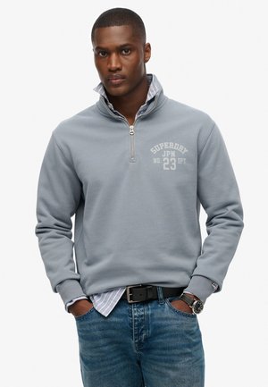 Sudadera gris con media cremallera y textura acanalada, con un logo blanco. Combinada sobre una camisa a rayas azules y blancas. Acompañada de unos vaqueros azules y un cinturón negro.