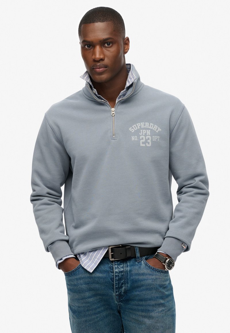 Sudadera gris con media cremallera y textura acanalada, con un logo blanco. Combinada sobre una camisa a rayas azules y blancas. Acompañada de unos vaqueros azules y un cinturón negro.