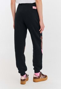 Osoba ubrana w czarne dresy z brązowymi i różowymi łatami, różowe skarpetki i brązowe trampki na platformie, stojąca na białym tle.