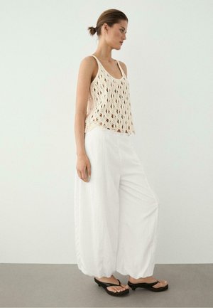 Vrouw die een beige gehaakte mouwloze top draagt, witte broek met wijde pijpen en zwarte sandalen met hakken, zijwaarts staand tegen een effen muur.