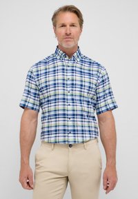 Camicia a maniche corte a quadri blu, verde e bianco; presenta bottoni e colletto; abbinata a pantaloni beige.