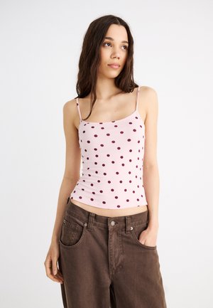 Νεαρή γυναίκα με μακριά σκούρα μαλλιά, ντυμένη με ροζ τοπ με πουά και άνετο καφέ παντελόνι, ποζάροντας με το ένα χέρι στην τσέπη.