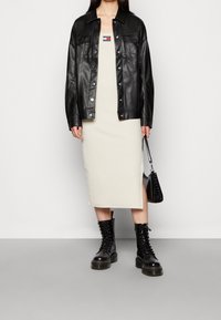 Černá kožená bunda přes krémové midi šaty s malým logem. Černé šněrovací armádní boty, držící texturovanou černou kabelku.