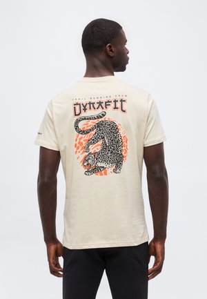 Hátát fordító férfi bézs pólóban, amelyen fekete és narancssárga, morajt mutató leopárd grafika és a „Trail Running Crew Dynafit” felirat található a hátán.