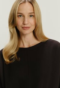 Blondynka o falowanych włosach, ubrana w ciemną bluzkę z okrągłym dekoltem, patrzy w przód z neutralnym wyrazem twarzy na jednolitym jasnym tle.