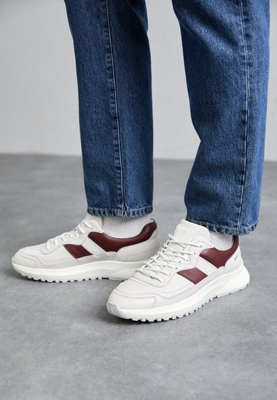 Λευκά και μπορντό αθλητικά παπούτσια με υφασμάτινη επένδυση από δέρμα, διαθέτοντας παχιά λευκή σόλα και αντίθετες ρίγες στα πλάγια.