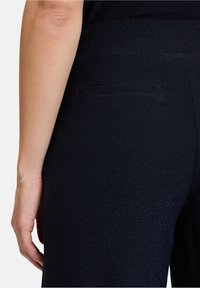 Schwarze Hosen mit einem dezenten Schimmer, die eine elegante, maßgeschneiderte Passform und eine einzelne Gesäßtasche aufweisen. Das Material wirkt strukturiert und glatt.