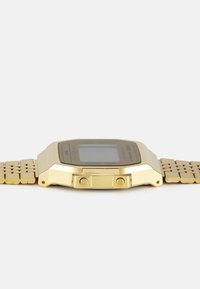 Montre numérique dorée avec un cadran rectangulaire, un bracelet en métal et deux boutons sur les côtés. L'affichage comporte un texte supplémentaire et une option de lampe de poche.