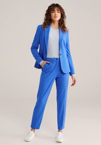 Plavi krojeni sako i odgovarajuće hlače od glatkog, rastezljivog materijala. Upareno s sivom majicom i bijelim tenisicama. Kroj ravnih nogavica.