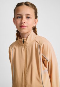  ferskenfarvet zip-up jakke med høj krave, struktureret stof og hvide accenter langs siderne. Har et lille logo på brystet.