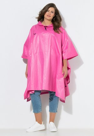 Donna che indossa un poncho impermeabile ampio di colore rosa acceso con cappuccio su jeans corti blu e sneakers bianche, in piedi davanti a uno sfondo bianco semplice.