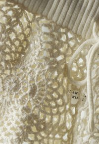 Cream-colored crochet garment with a textured pattern, featuring a ribbed neckline and drawstring detail for adjustment.