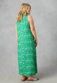 Grön ärmlös maxiklänning med blommönster, lång, flödande design, sidoflik och en texturerad tygkvalitet. Bärs med platta sandaler.