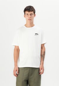 White cotton t-shirt with a round neckline, short sleeves, and a small black graphic print on the chest. Model paired with olive green pants.
