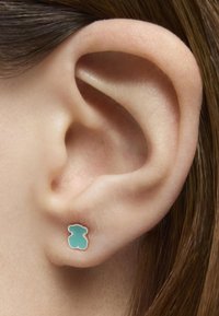 Pendiente de perrito de goma turquesa con acabado suave, contorneado en metal tono oro, llevado en la oreja de un modelo. Diseño simple y juguetón.
