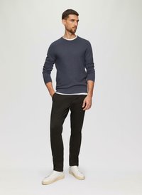 Marineblaues Strickpullover mit Rundhalsausschnitt, darüber ein weißes Hemd. Schwarze schmal geschnittene Hose und weiße Sneakers runden das Outfit ab.