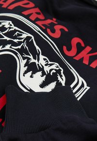 Marineblauer Sweatshirt mit einem großen weißen und roten Grafikdesign, das eine Bergkontur zeigt, gerippten Bündchen und einer weichen Stofftextur.