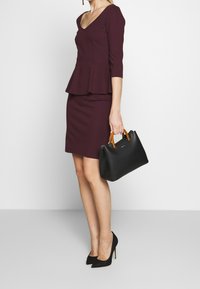 Burgundy peplumtröja med trekvartsärmar och matchande figursydd kjol. Svart handväska med bruna handtag. Svarta stilettohöljar.