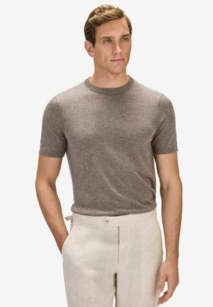 Man met kort lichtbruin haar draagt een nauwsluitend kort mouw bruin gebreid shirt en lichtbeige hoge taille broek, staand met één hand in de zak.