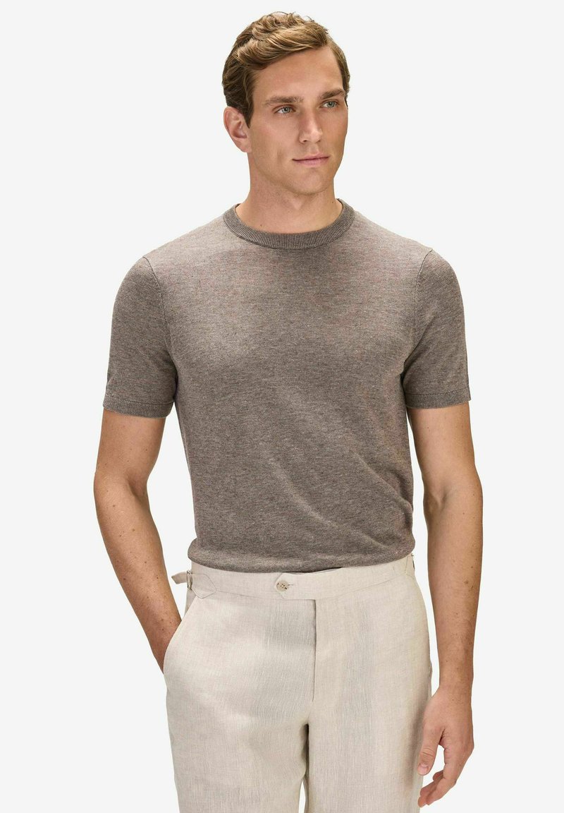 Homme aux cheveux courts châtain clair portant un pull en maille marron à manches courtes ajusté et un pantalon taille haute beige clair, debout avec une main dans la poche.