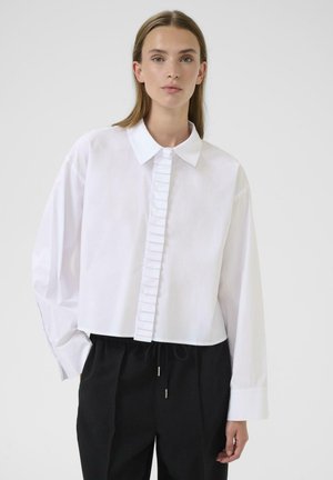 Camicia bianca cropped con colletto strutturato e placca a rouches verticale. Maniche lunghe con una consistenza liscia. Indossata con pantaloni neri.