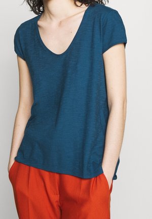 Osoba nosząca morski zielony t-shirt z krótkim rękawem i dekoltem w serek oraz jasnopomarańczowe spodnie, ręce w kieszeniach, na neutralnym tle.