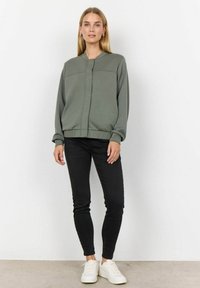 Světle zelená bomber bunda s žebrovanými manžetami a lemem, se zapínáním na přední zip; nošena s úzkými černými džíny a bílými teniskami.