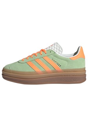 Niskie buty Adidas Gazelle w jasnym zielonym zamszu z pomarańczowymi paskami, sznurowadłami, tylnym panelem i potrójnie warstwową brązową podeszwą.