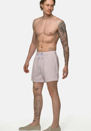 Man zonder shirt met mouwtattoo op de rechterarm en volledige tattoo op het linkerbeen, draagt lichtgekleurde korte broek en instapschoenen, staat en kijkt naar links.