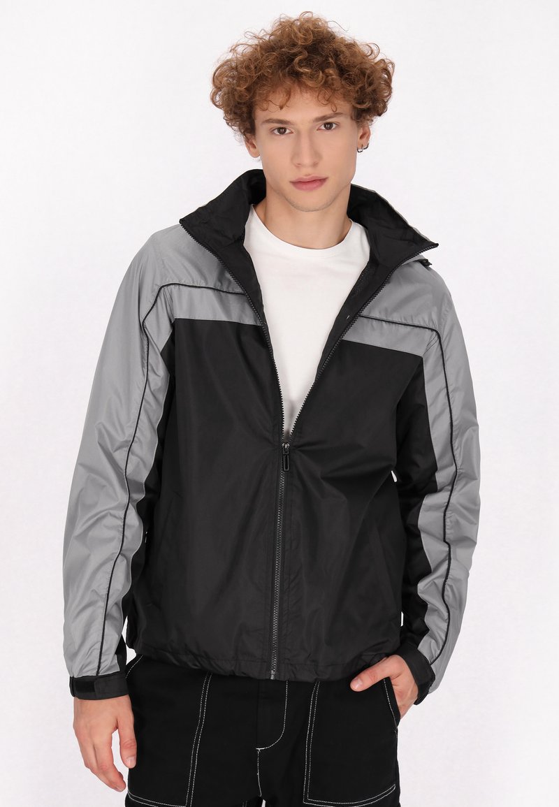 Chaqueta cortavientos negra y gris con cierre de cremallera en la parte delantera, cuello ajustable y paneles laterales en contraste. Textura suave con puños elásticos.