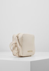 Bežová kožená crossbody taška se zlatým zipem, zlatými kovovými prvky a vyraženým názvem značky. Kompaktní obdélníkový tvar. Texturovaný povrch.